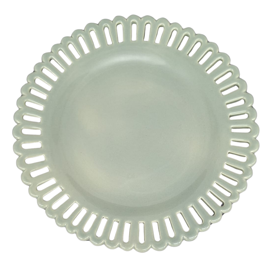 A sage green round ceramic cake plate on a low foot with a scalloped, openwork rim featuring elongated slit-like cutouts, shown from a top-down view on a white background.